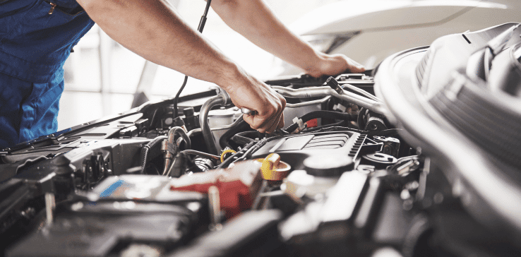 A mechanic wearing a blue uniform is working on the engine of a car. The car's hood is open, and the mechanic's hands are visible as they handle tools and components inside the engine bay. The scene is well-lit, showcasing various parts of the car's engine.